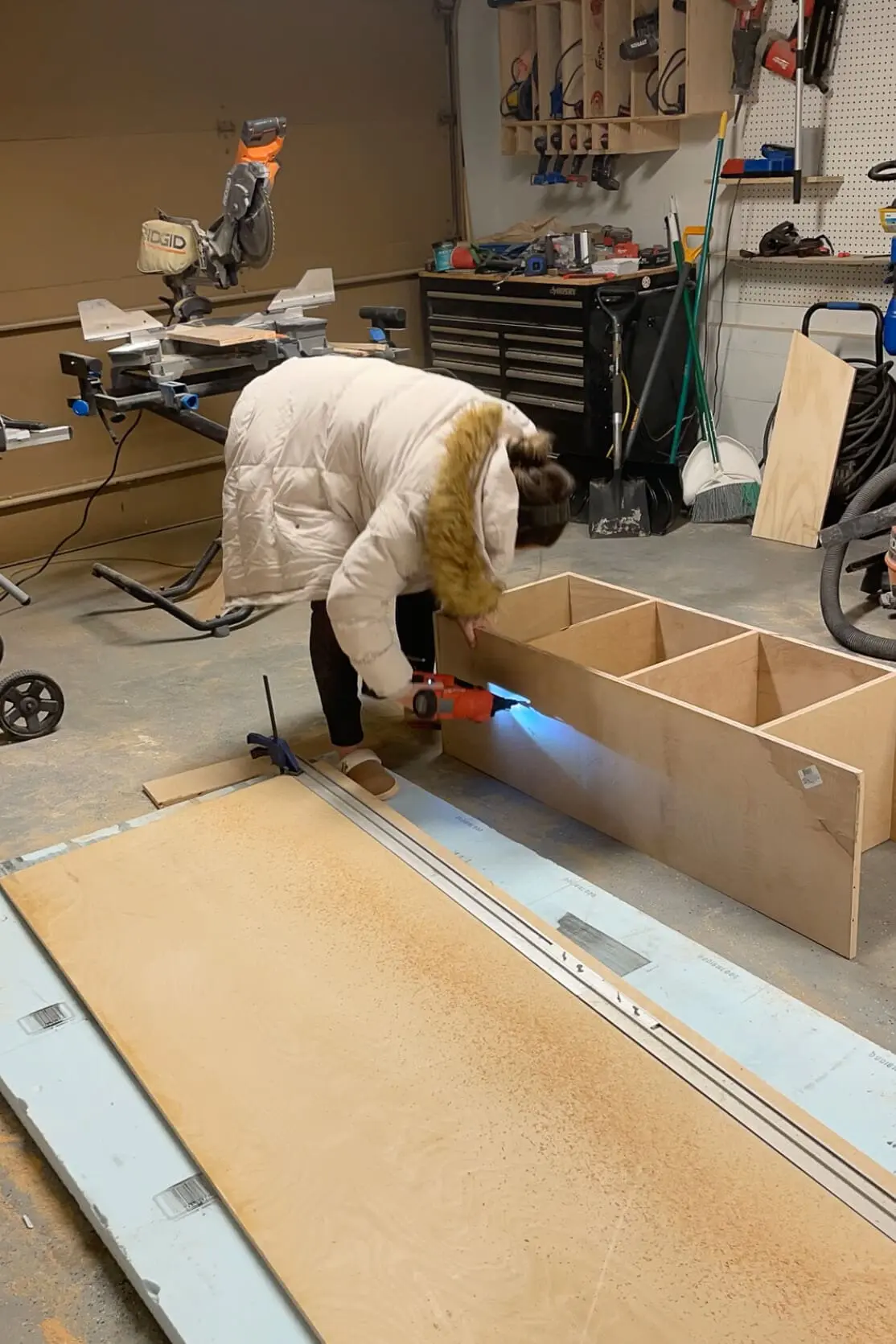 Woman in a warm winter coat assembling a wooden cabinet in a workshop. She uses a cordless nail gun to secure the front panel while working on a plywood workbench surrounded by tools, including a miter saw and clamps.
